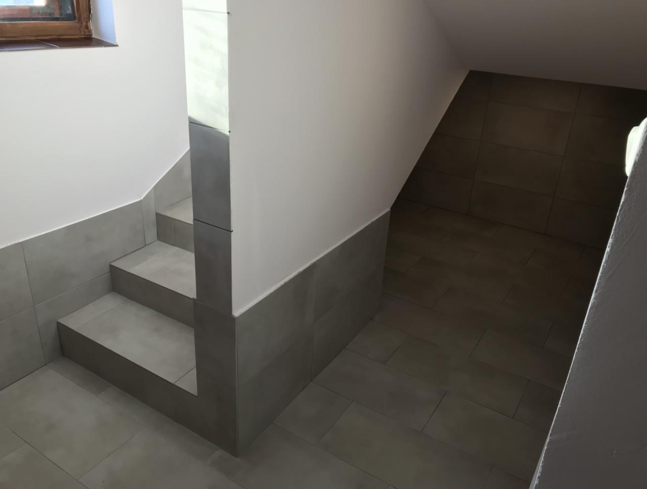 Wykończone schody z szarymi płytkami ceramicznymi, prowadzące w dół, z widocznym fragmentem okna w górnym lewym rogu.