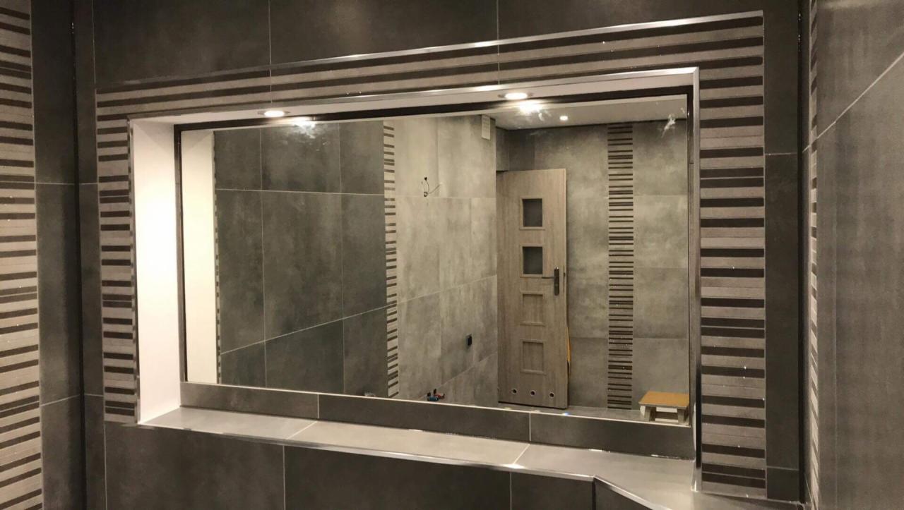 Wykończona łazienka z szarą glazurą, ozdobnymi pasami płytek i lustrem w ramie z płytek. Widoczna drewniana łazienkowa szafka pod umywalką.