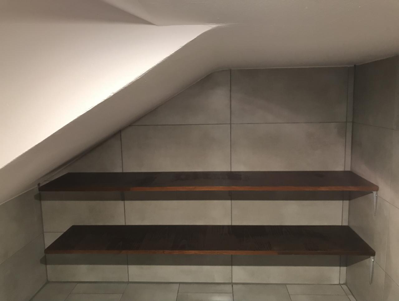 Ukośna ściana wyłożona szarymi płytkami ceramicznymi, dwie ciemnobrązowe półki zamontowane na metalowych wspornikach, na dole szare płytki podłogowe.