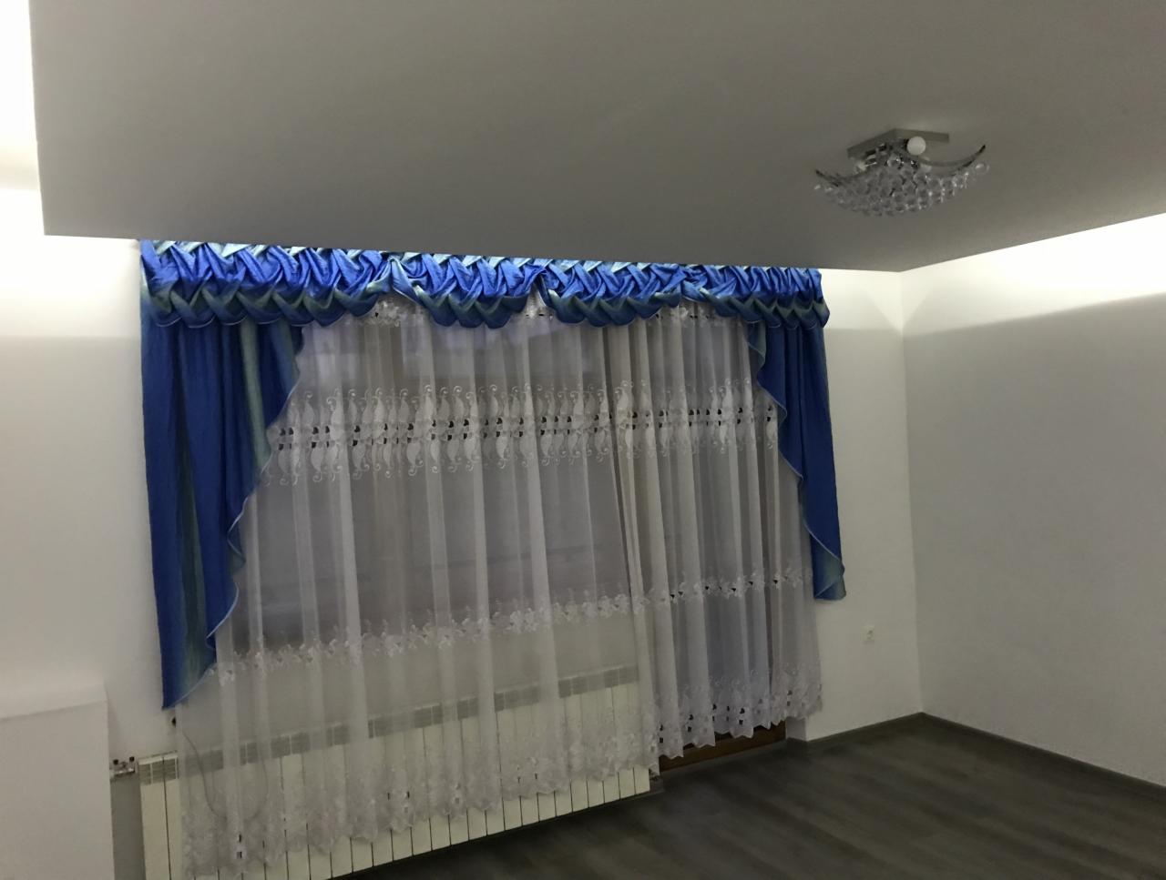 Okno z założonymi, długimi, białymi firanami z delikatnym haftem i niebieskimi zasłonami z ozdobną falbaną, widoczny grzejnik i szara podłoga.