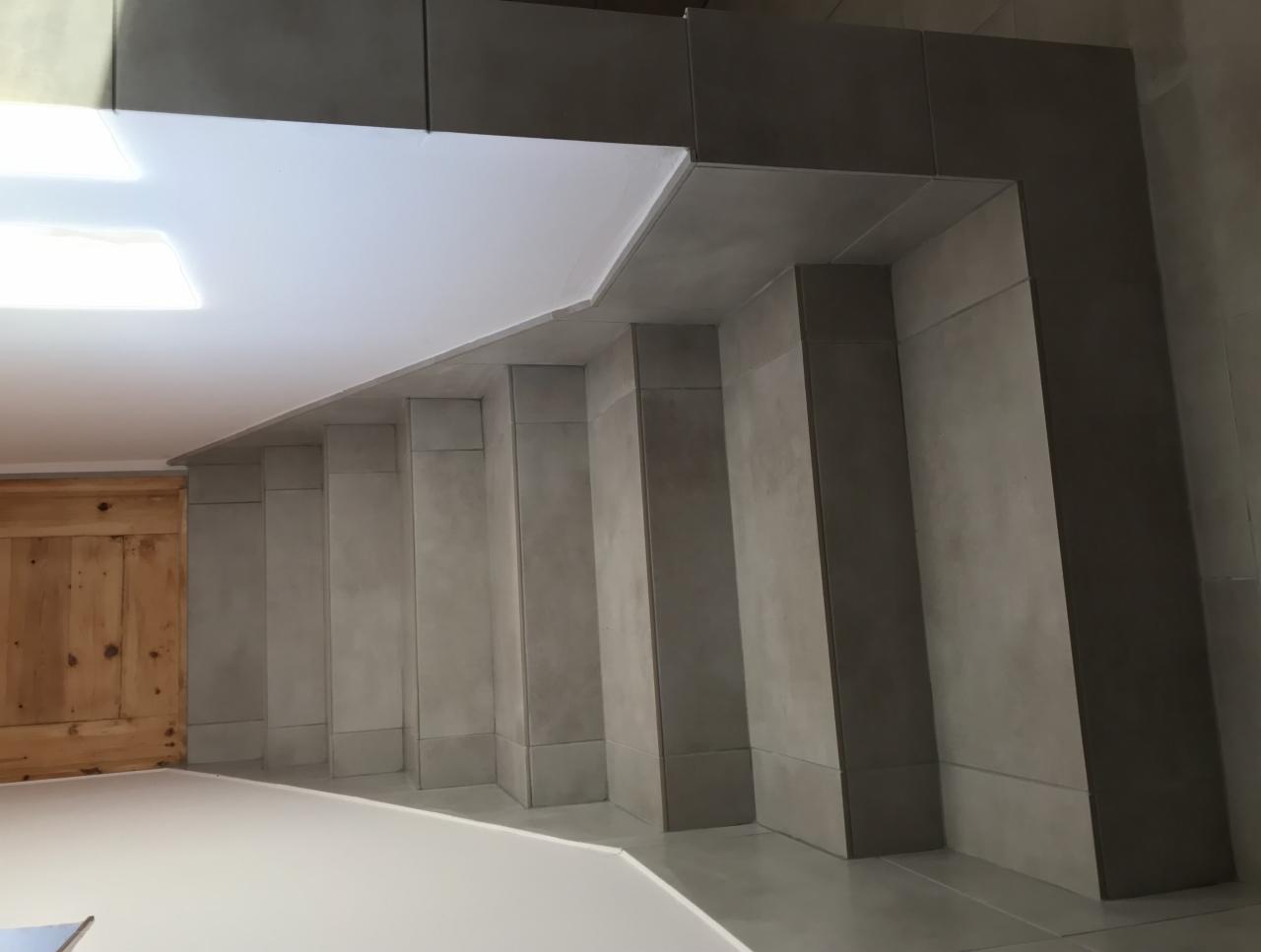 Wykończone wnętrze z szarymi, matowymi płytkami na ścianach i podłodze, widoczne narożniki i krawędzie płytek, biały sufit i drewniane drzwi w tle.