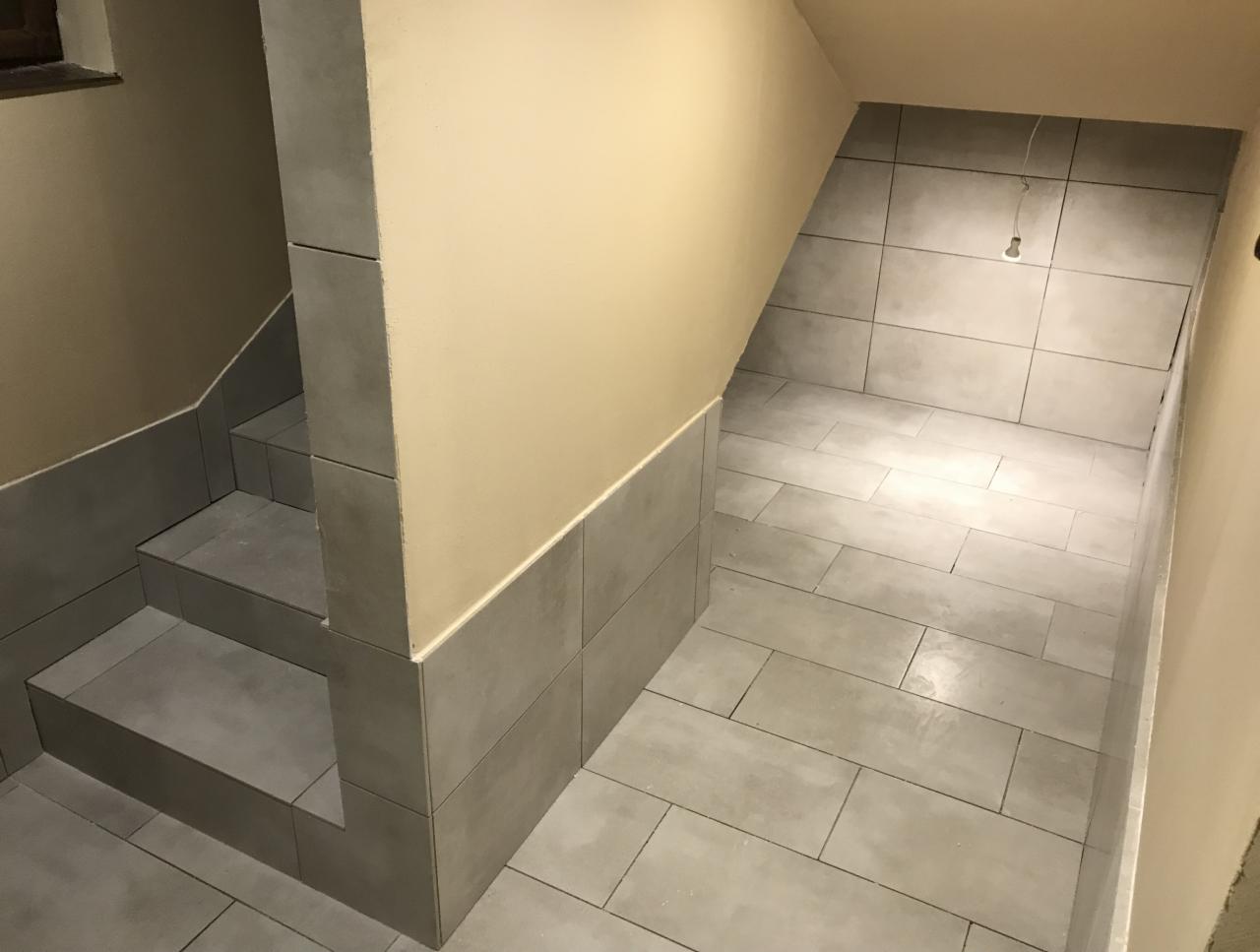 Wykończone płytkami schody prowadzące w dół, z widocznymi szarymi płytkami na stopniach, ścianach i podłodze, oświetlone wiszącą żarówką bez klosza.