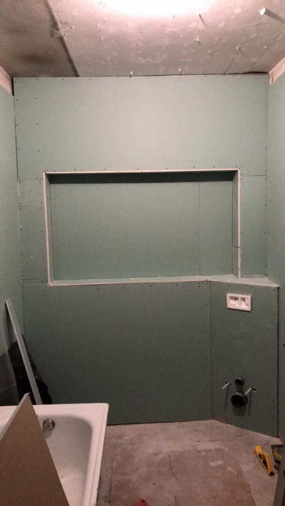 Łazienka w trakcie remontu: ściany wyłożone zieloną płytą kartonowo-gipsową z wnęką, widoczne rury instalacji wodnej, fragment wanny i poziomicy na podłodze.