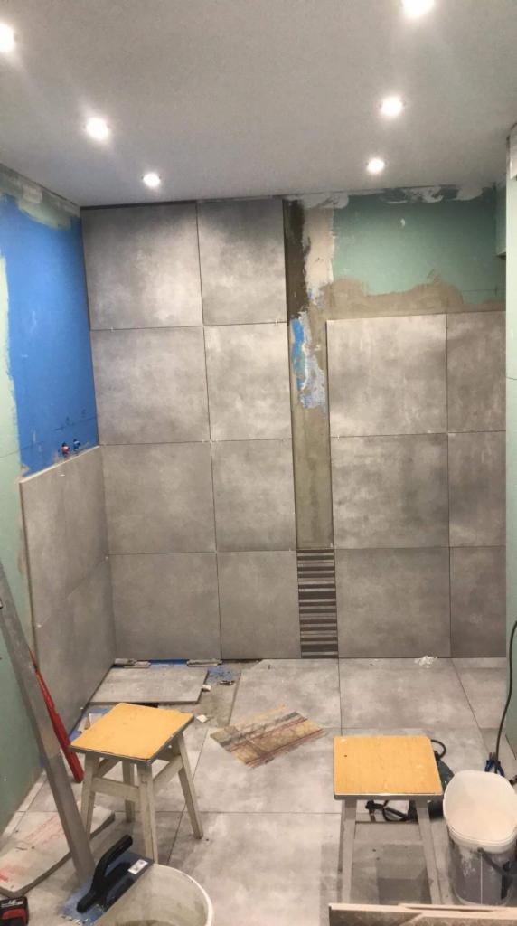 Łazienka w trakcie wykańczania: szare, kwadratowe płytki częściowo ułożone na ścianach i podłodze, widoczne narzędzia, drabina i dwa stołki.