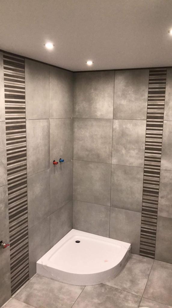 Wykończona łazienka z szarymi, kwadratowymi płytkami, dekoracyjnymi pasami z poziomych pasków i białą, narożną kabiną prysznicową.