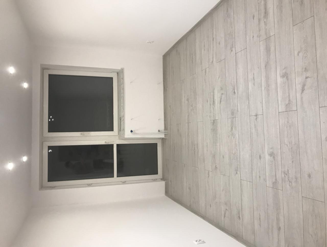 Pokój po remoncie: ściana wykończona panelami ściennymi imitującymi jasne drewno, białe ściany, okno dwukomorowe z białymi ramami, widok z dołu.