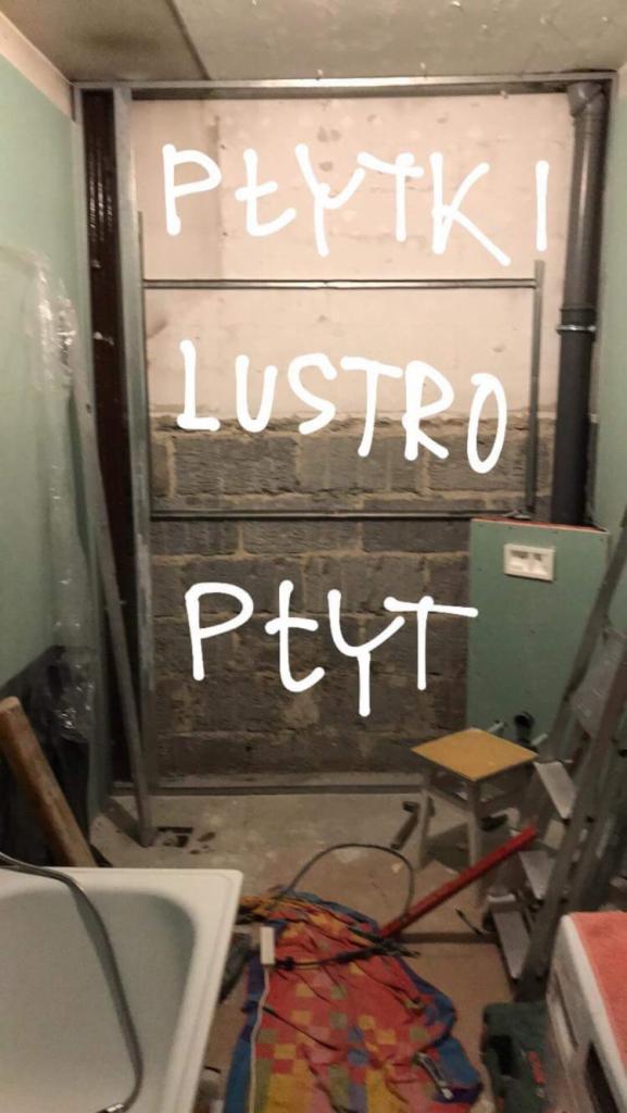 Przygotowania do remontu łazienki: widoczne stelaże pod zabudowę, fragmenty instalacji, narzędzia i materiały budowlane w nieładzie, ściany z surowej cegły z napisanymi odręcznie słowami: 'Płytki',...