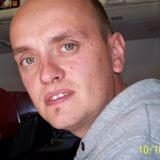 Portret mężczyzny z bliska, ubrany w szarą bluzę z kapturem, z widocznym znamieniem na brodzie, na tle jasnej ściany.