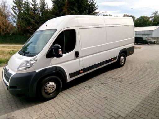 Biały bus dostawczy Citroen Jumper z czarnymi elementami wykończenia, zaparkowany na kostce brukowej, widok z przodu i boku, zarys budynku w tle.