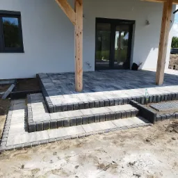 Nowo ułożona, szara kostka brukowa tworząca taras i schody przy domu z drewnianą kolumną podtrzymującą zadaszenie, widoczne niedokończone prace ziemne.