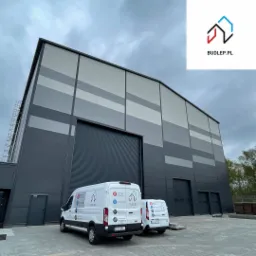 Duży, szary budynek przemysłowy z dwoma białymi samochodami dostawczymi z logo firmy BUDLEP.PL, świadczącej usługi instalacyjne, klimatyzacyjne i budowlane.