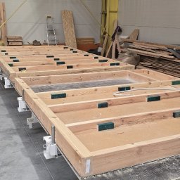 BUDLEP SPÓŁKA Z OGRANICZONĄ ODPOWIEDZIALNOŚCIĄ - Wnętrze hali z drewnianymi ramami ścian szkieletowych w trakcie prefabrykacji, widoczne podpisy pomieszczeń, drabina w tle. Elementy domu szkieletowego na etapie produkcji.