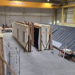 BUDLEP SPÓŁKA Z OGRANICZONĄ ODPOWIEDZIALNOŚCIĄ - Wnętrze hali z elementami domu szkieletowego w trakcie prefabrykacji. Widoczna konstrukcja ścian z oknami, dach z papy, rusztowanie i dźwig Konecranes.