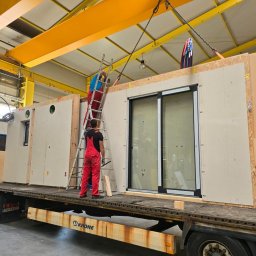 BUDLEP SPÓŁKA Z OGRANICZONĄ ODPOWIEDZIALNOŚCIĄ - Montaż modułowego domu szkieletowego na naczepie ciężarówki. Dwóch monterów w czerwonych kombinezonach pracuje przy użyciu drabiny i dźwigu. Widoczne okno przesuwne.