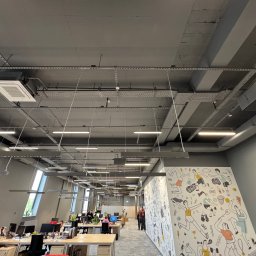 BUDLEP SPÓŁKA Z OGRANICZONĄ ODPOWIEDZIALNOŚCIĄ - Klimatyzacja kasetonowa w nowoczesnym biurze z widocznymi instalacjami pod stropem. Oświetlenie liniowe i dekoracyjna ściana z rysunkami sportowymi.