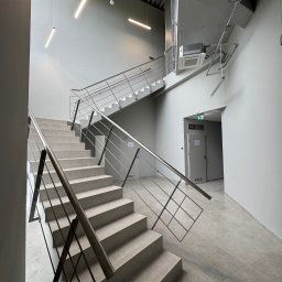 BUDLEP SPÓŁKA Z OGRANICZONĄ ODPOWIEDZIALNOŚCIĄ - Betonowe schody z metalową balustradą w nowoczesnym, minimalistycznym wnętrzu z szarą kolorystyką i oświetleniem liniowym. Widoczny klimatyzator sufitowy.