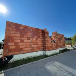 Wznoszenie ściany z czerwonej cegły na fundamencie z izolacją, widok z zewnątrz na tle błękitnego nieba i częściowo zadaszonego domu w budowie.