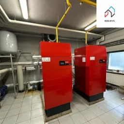 Dwa czerwone piece gazowe Hoval UltraGas w kotłowni z białymi rurami i żółtymi przewodami gazowymi, jasne płytki podłogowe, widoczne elementy instalacji.