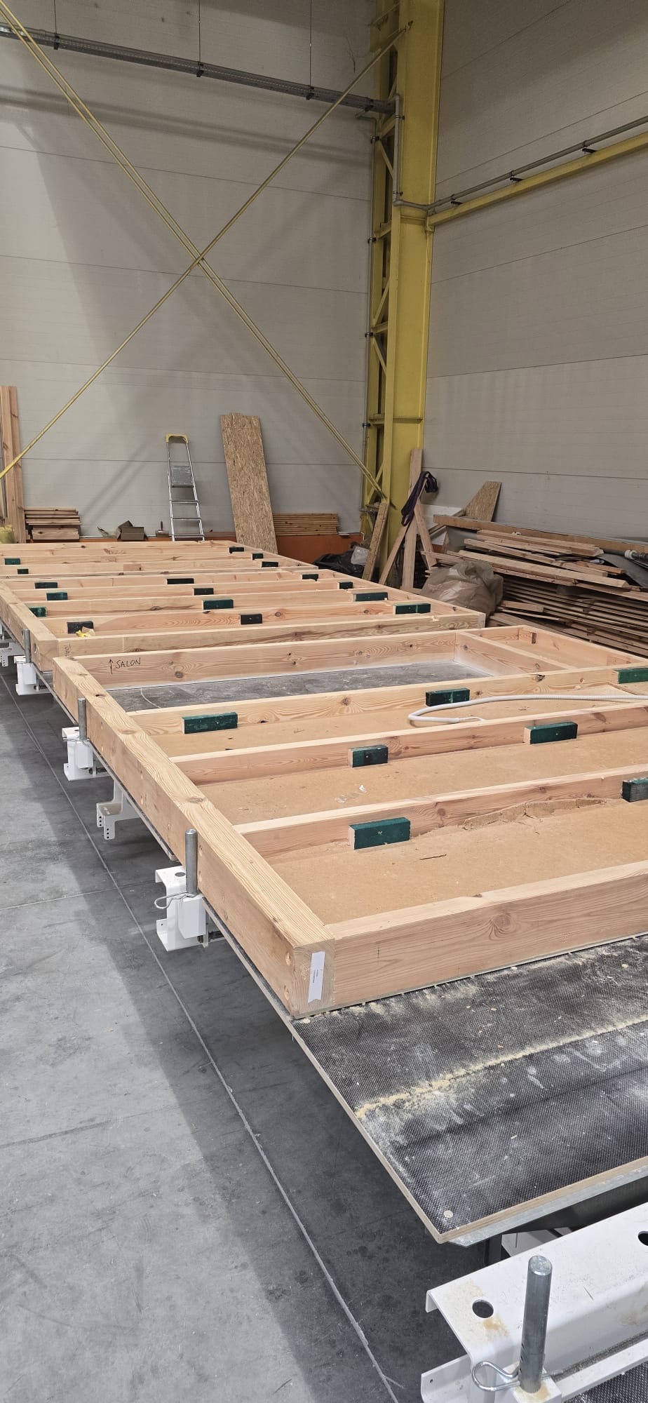 Elementy konstrukcyjne ścian szkieletowych w hali produkcyjnej, widoczne drewniane ramy z oznaczeniem 'SALON', mocowane na metalowych wspornikach, w tle drabina i składowane deski.