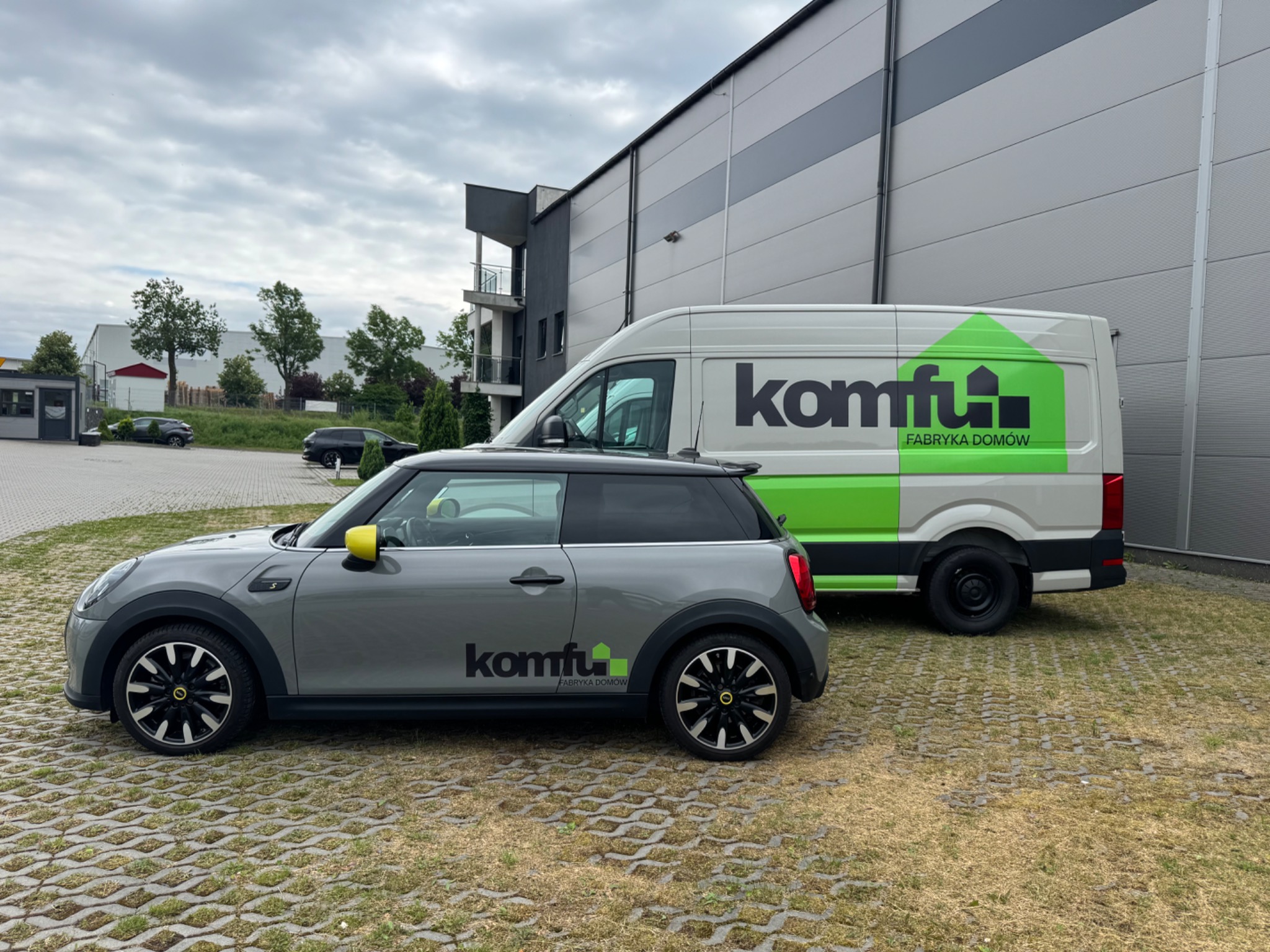 Szary Mini Cooper i biały van z logo firmy budującej domy 'Komfu Fabryka Domów' na tle szarego budynku. Kąt ujęcia z boku, pochmurne niebo.