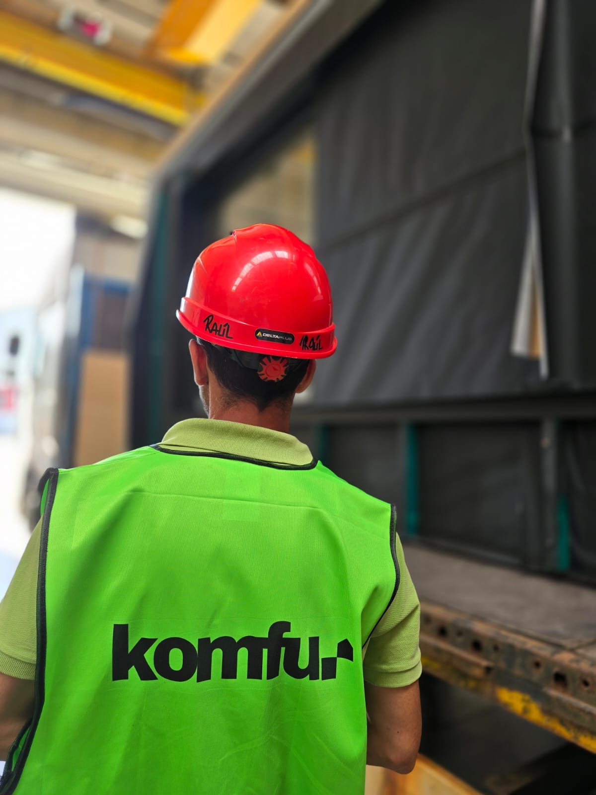 Pracownik w kasku i kamizelce z logo 'komfur' stoi tyłem do kamery, w tle widoczna naczepa ciężarówki i elementy konstrukcji hali.