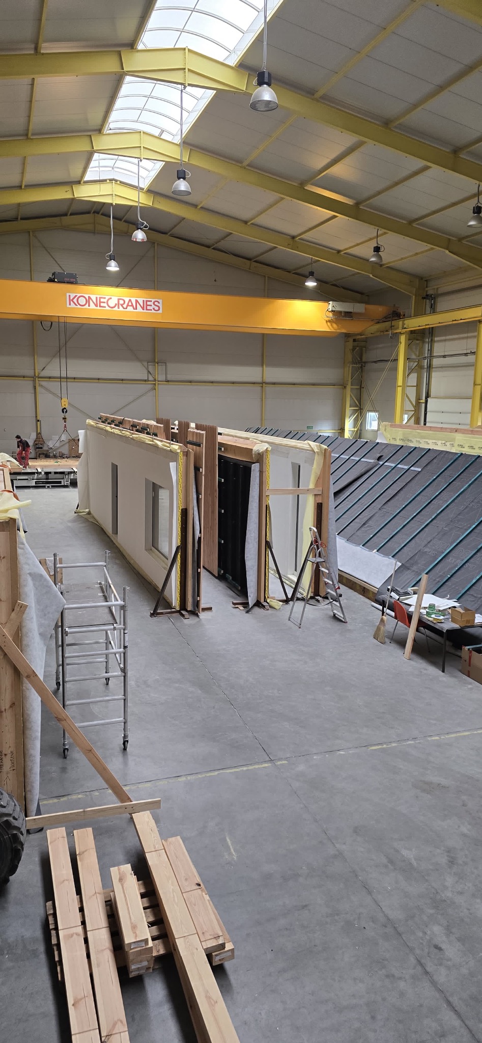 Wnętrze hali z elementami domu szkieletowego w trakcie prefabrykacji. Widoczna konstrukcja ścian z oknami, dach z papy, rusztowanie i dźwig Konecranes.