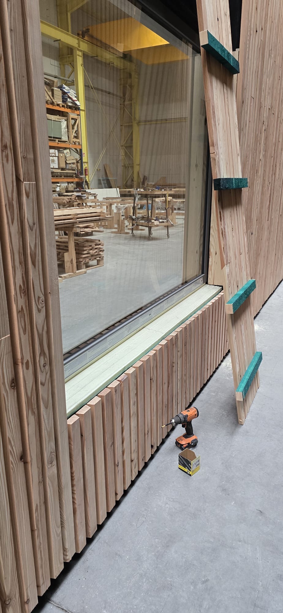Montaż drewnianej elewacji z oknem, widoczna wiertarka i pudełko wkrętów. Elementy elewacji z zielonymi akcentami. Hala produkcyjna w tle.