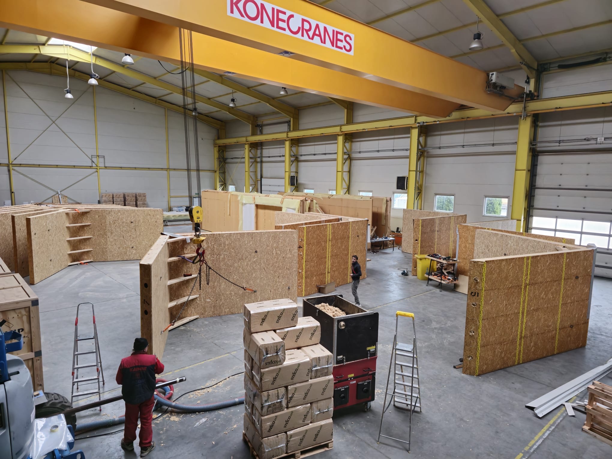 Montaż ścian domu szkieletowego w hali produkcyjnej z użyciem suwnicy Konecranes. Widoczne elementy konstrukcyjne, materiały izolacyjne Steico i pracownicy.