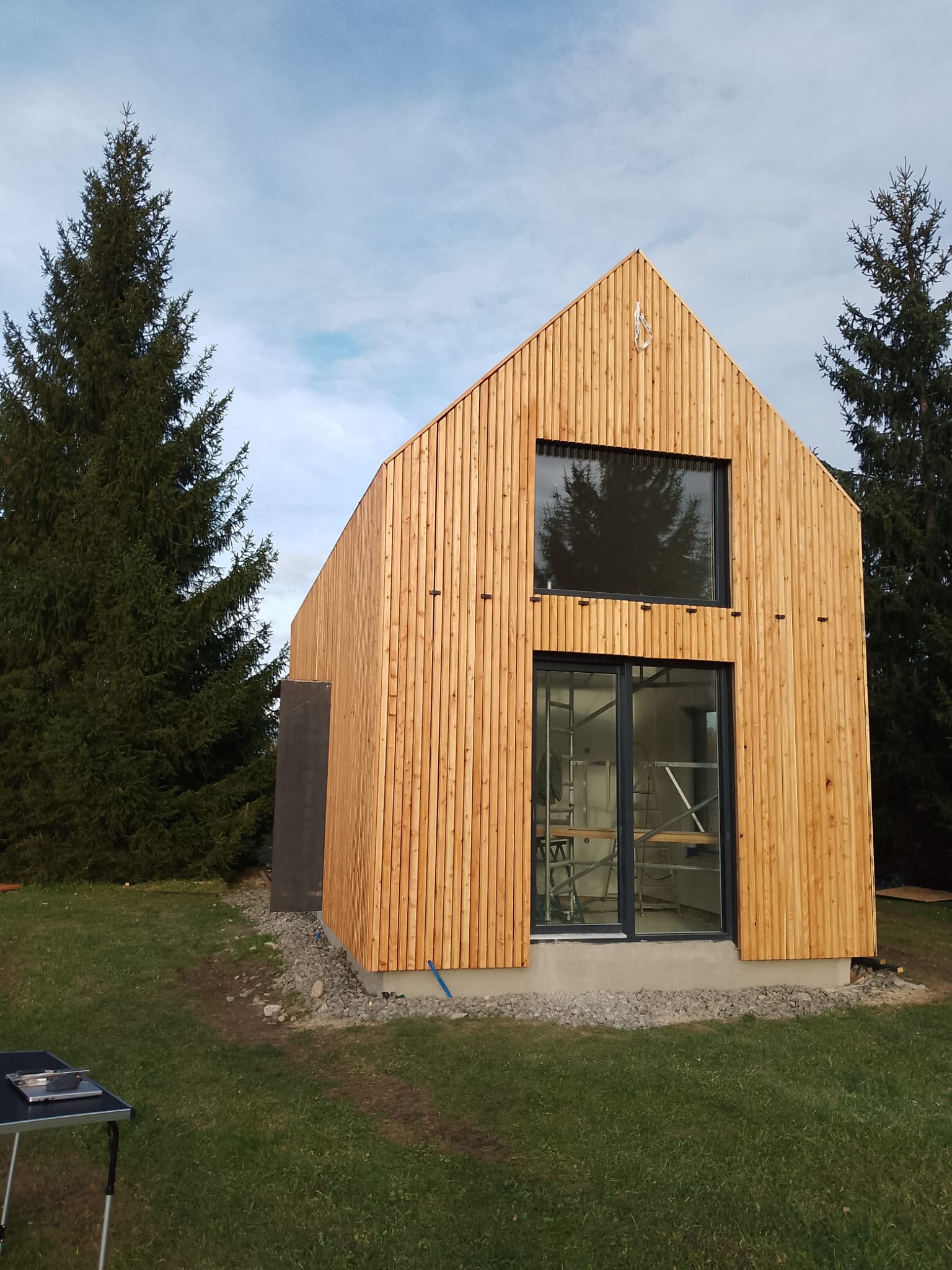 Nowoczesny dom szkieletowy z drewnianą elewacją w pionowym układzie desek. Widoczne okna z czarnymi ramami i rusztowania wewnątrz. Dom otoczony zielenią.