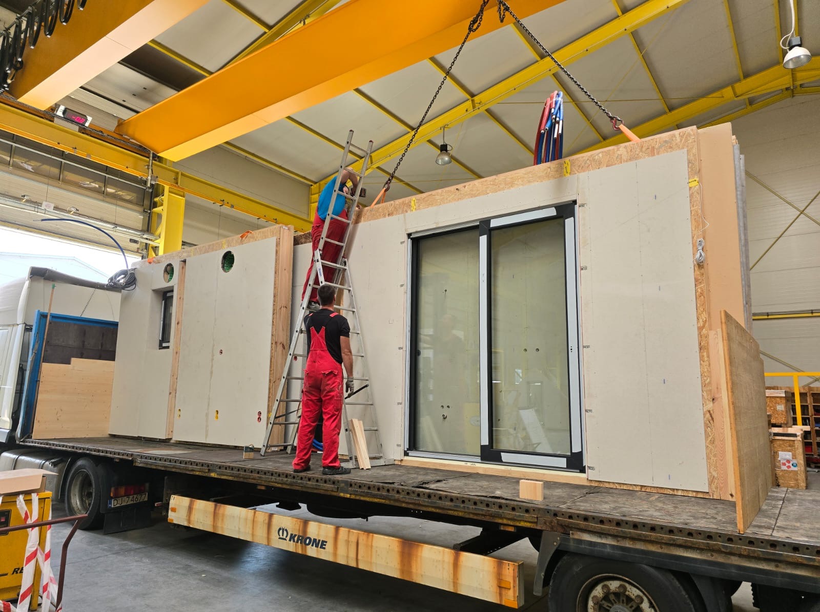 Montaż modułowego domu szkieletowego na naczepie ciężarówki. Dwóch monterów w czerwonych kombinezonach pracuje przy użyciu drabiny i dźwigu. Widoczne okno przesuwne.