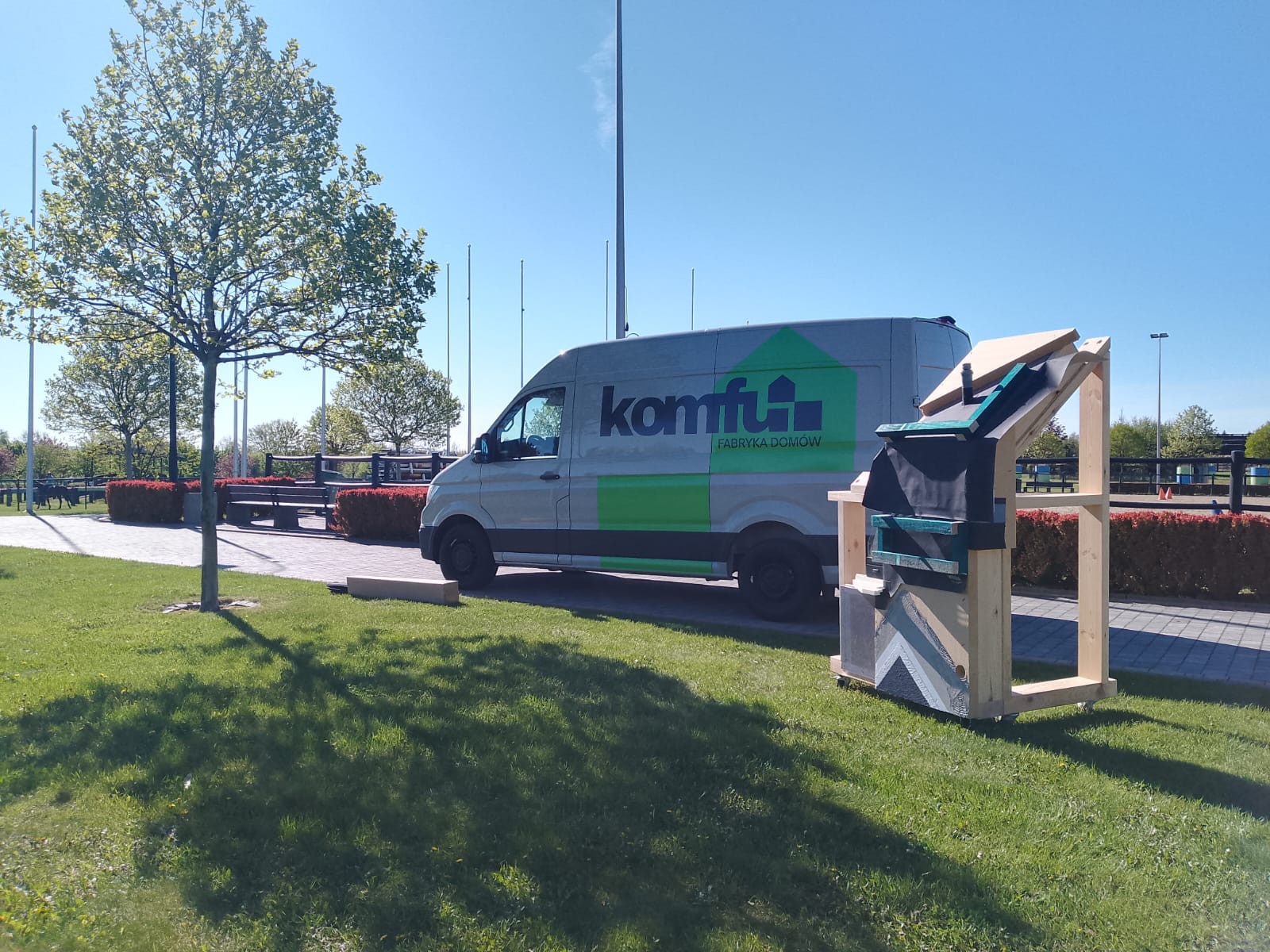 Prezentacja przekroju dachu z izolacją i pokryciem, obok samochód firmowy 'Komfu Fabryka Domów' na trawniku w słoneczny dzień. Model edukacyjny.