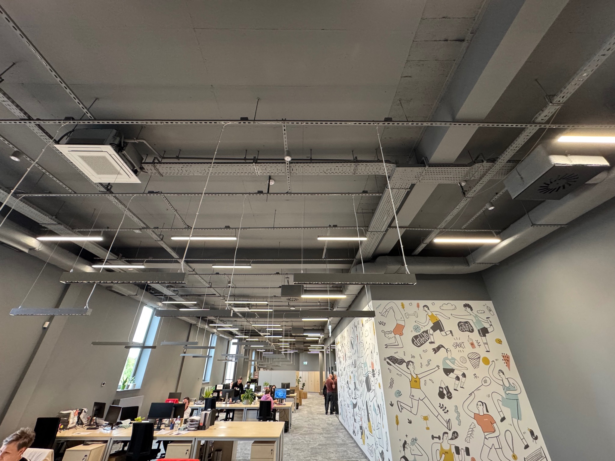 Klimatyzacja kasetonowa w nowoczesnym biurze z widocznymi instalacjami pod stropem. Oświetlenie liniowe i dekoracyjna ściana z rysunkami sportowymi.