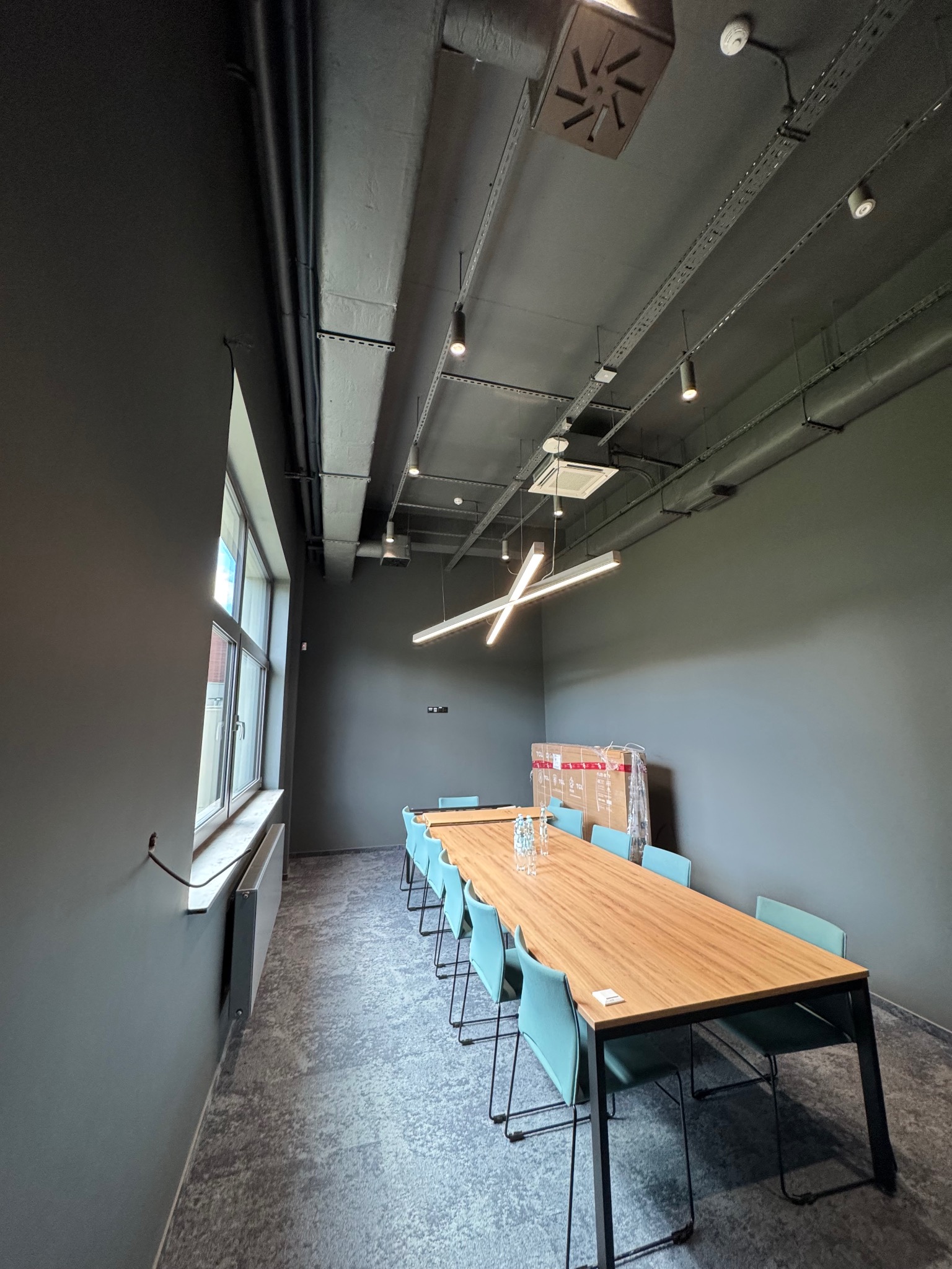 Nowoczesna sala konferencyjna z długim stołem, krzesłami w kolorze morskim i minimalistycznym oświetleniem sufitowym. Widoczne instalacje pod sufitem i grzejnik pod oknem.