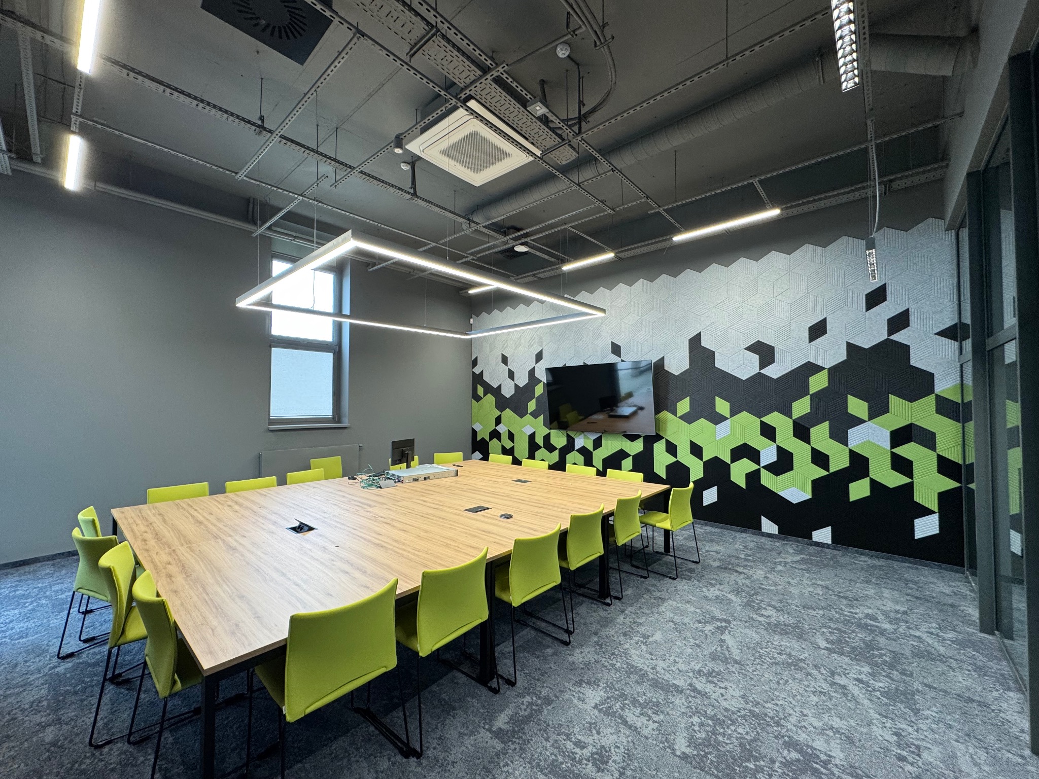 Nowoczesna sala konferencyjna z długim stołem, zielonymi krzesłami i dekoracyjną ścianą w geometryczne wzory. Widoczna klimatyzacja kasetonowa pod sufitem.