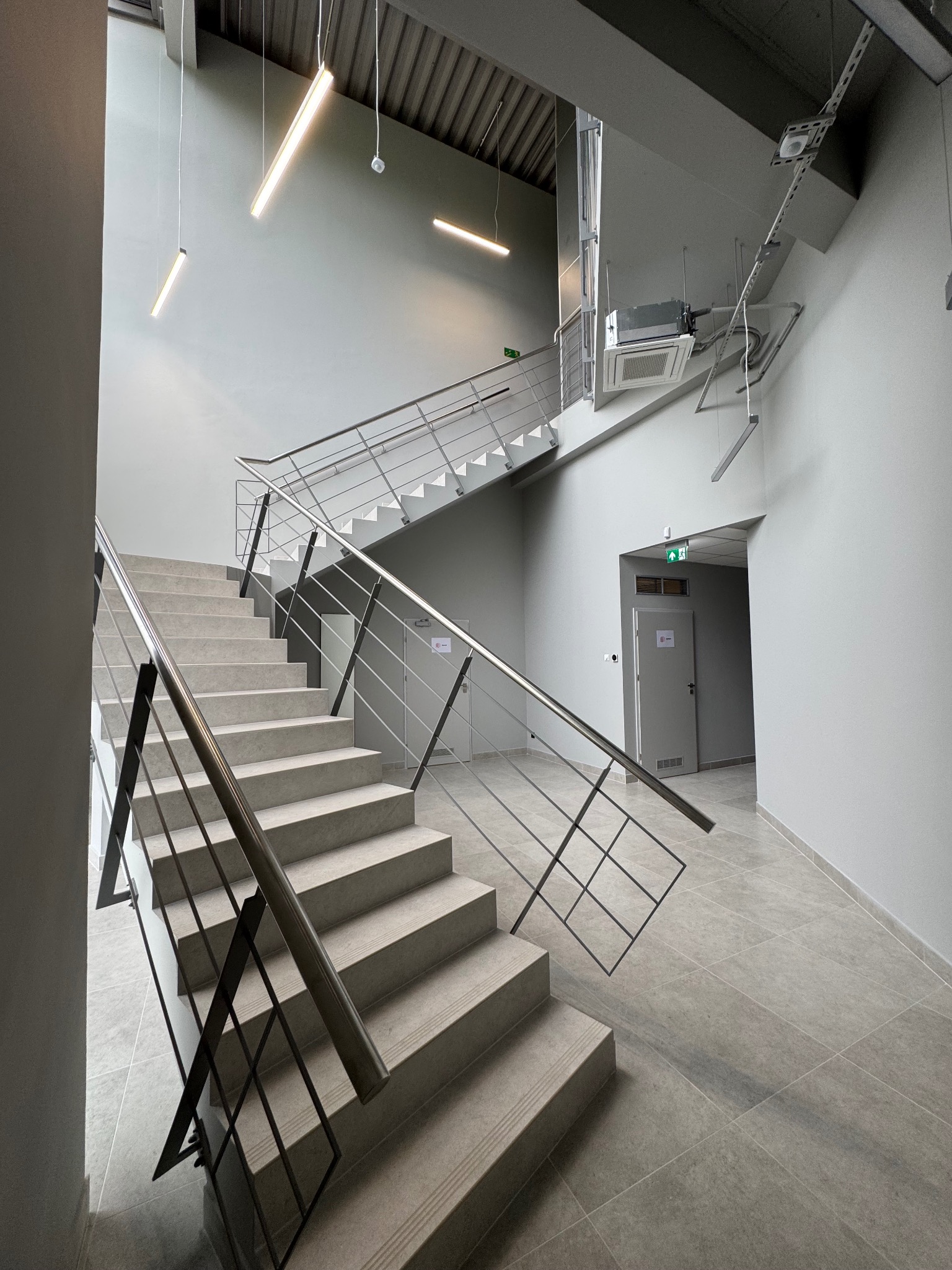 Betonowe schody z metalową balustradą w nowoczesnym, minimalistycznym wnętrzu z szarą kolorystyką i oświetleniem liniowym. Widoczny klimatyzator sufitowy.