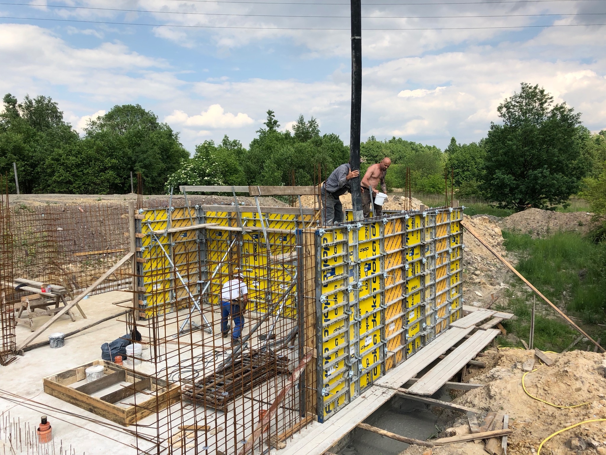 Budowa fundamentów: Dwóch robotników wylewa beton z pompy w szalunki, trzeci pracuje wewnątrz konstrukcji. Wokół zbrojenia i drewniane elementy. Tło: zieleń drzew i błękitne niebo.