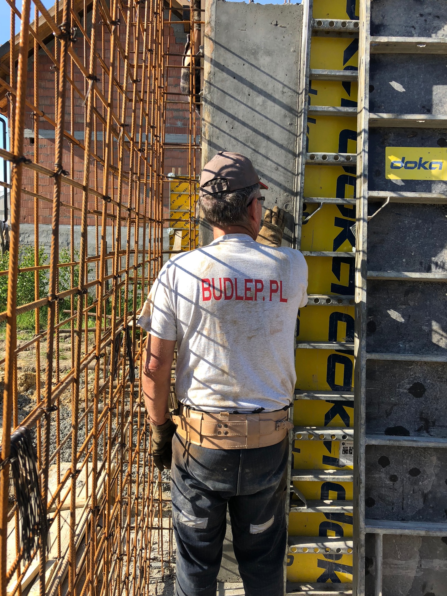 Pracownik w trakcie montażu szalunków i zbrojenia na budowie domu murowanego, widok z tyłu, na koszulce napis BUDLER.PL. Elementy konstrukcyjne Doka.