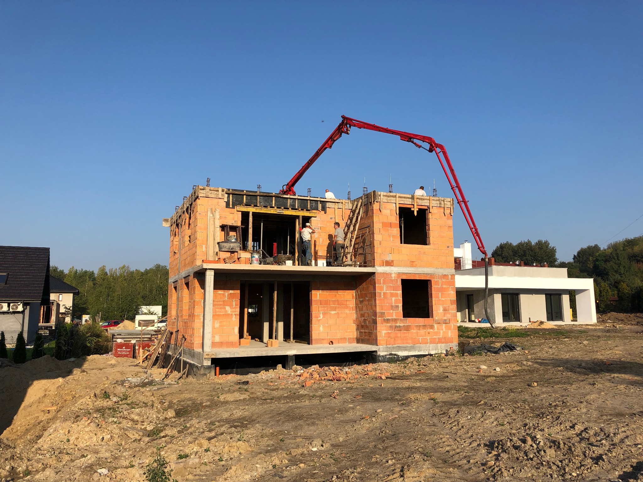 Budowa domu murowanego w trakcie wznoszenia ścian z czerwonej cegły. Na górze pracownicy obsługują czerwoną pompę do betonu. W tle widoczny inny budynek i drzewa.