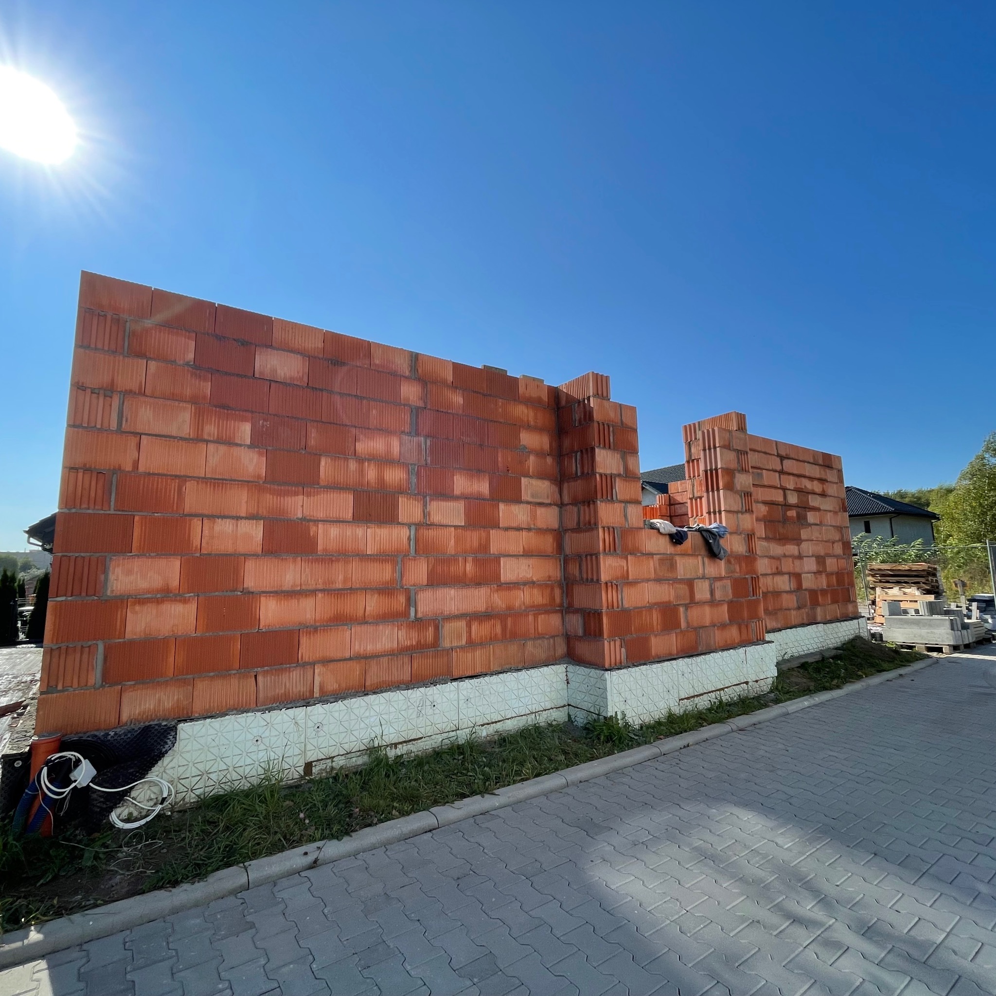 Wznoszenie ściany z czerwonej cegły na fundamencie z izolacją, widok z zewnątrz na tle błękitnego nieba i częściowo zadaszonego domu w budowie.