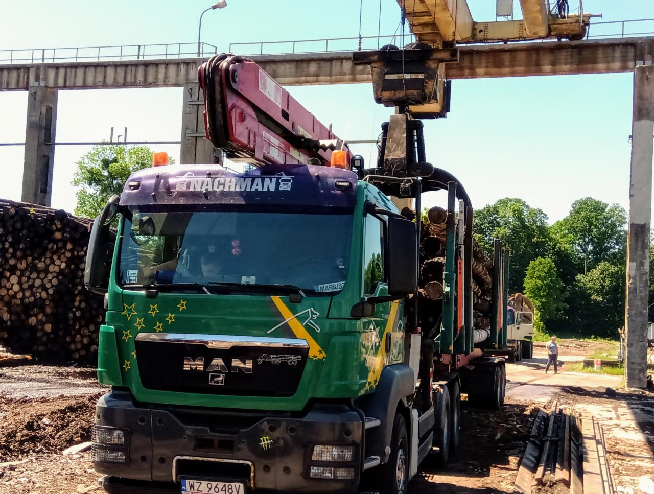 Ciężarówka MAN z dźwigiem załadowana drewnem na placu składowym, widoczny numer rejestracyjny i logo firmy.