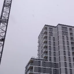 Budynek wielorodzinny w trakcie budowy, widziany z dołu w zimowy dzień. Na pierwszym planie żuraw budowlany i elementy konstrukcyjne.