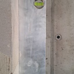 Pol-Bud Professional - Pionowy poziomica na surowej ścianie z betonu komórkowego, kontrola pionu, widoczne ślady po szpachlowaniu. Szary odcień betonu.