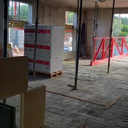 Pol-Bud Professional - Wnętrze surowego budynku w trakcie budowy. Widoczne palety z płytami gipsowymi MultiGips, podpory stropowe i fragment rusztowania. Surowy beton i naturalne światło.