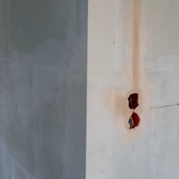 Pol-Bud Professional - Narożnik ścian po szpachlowaniu, widoczne puszki elektryczne z przewodami. Ściana po lewej w szarym odcieniu, po prawej jaśniejsza, przygotowana pod malowanie.