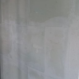 Pol-Bud Professional - Ściana z szarej płyty kartonowo-gipsowej, częściowo wyszpachlowana, z widocznymi śladami po szpachlowaniu i numerem '22' napisanym na powierzchni. U dołu widoczne gruz.