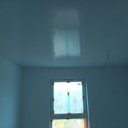 Pol-Bud Professional - Wnętrze pokoju po świeżym malowaniu na biało, z oknem zabezpieczonym folią. Widoczne refleksy światła na gładkiej powierzchni sufitu i ścian.