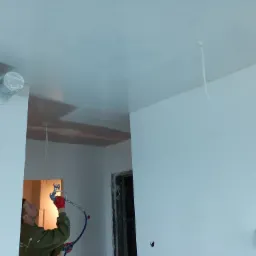 Pracownik w czerwonych rękawicach natryskowo nanosi białą farbę na sufit w pomieszczeniu. Widoczne zabezpieczone folią rury wentylacyjne i fragment ściany.