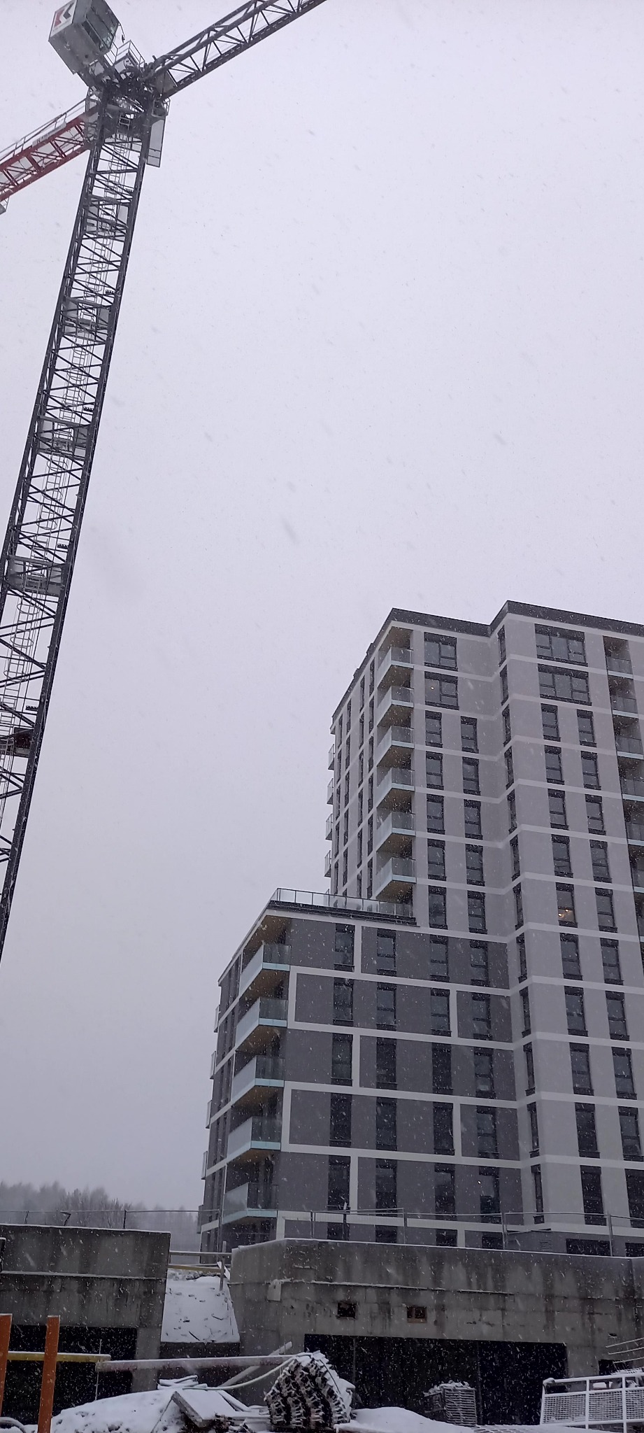Budynek wielorodzinny w trakcie budowy, widziany z dołu w zimowy dzień. Na pierwszym planie żuraw budowlany i elementy konstrukcyjne.