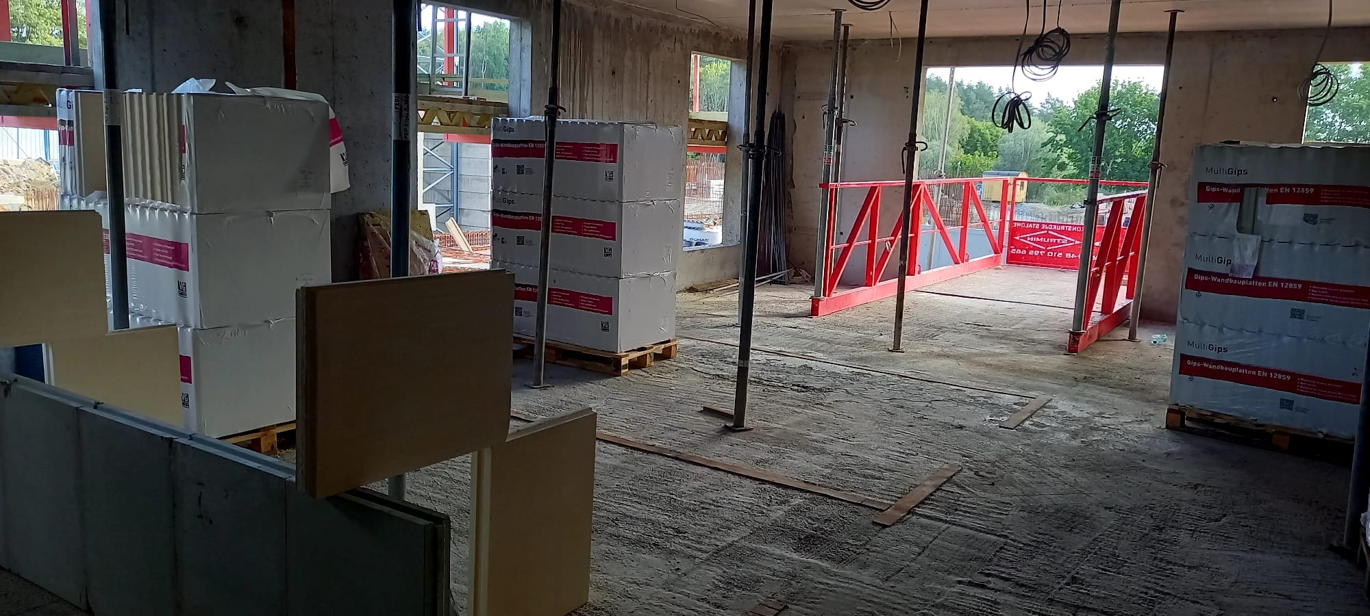 Wnętrze surowego budynku w trakcie budowy. Widoczne palety z płytami gipsowymi MultiGips, podpory stropowe i fragment rusztowania. Surowy beton i naturalne światło.