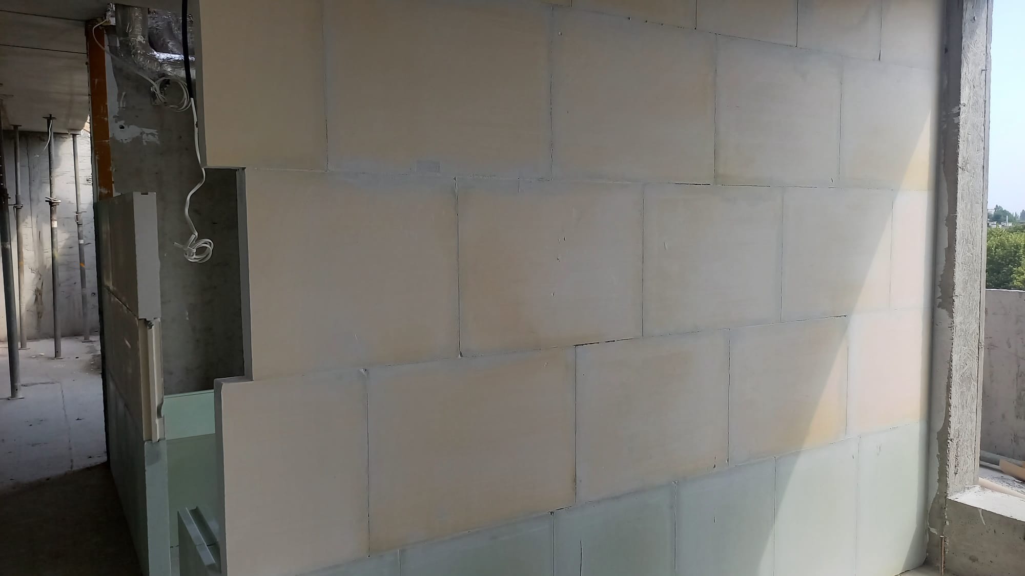 Wnętrze w trakcie budowy: ściany wyłożone płytami, widoczne instalacje i surowy beton. Jasne, naturalne światło podkreśla strukturę materiałów.